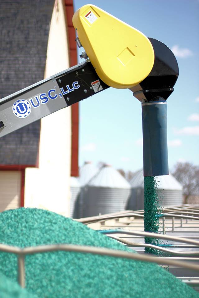 Automated grain feeder delivering green feed into a trough, with silos in the background. This image highlights agricultural machinery in use.